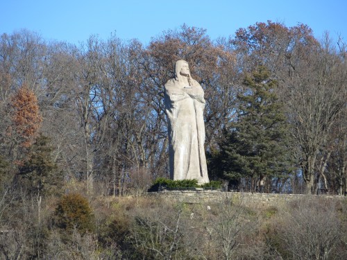 A large statue of a figure standing on a hillside, surrounded by trees and a clear blue sky.
