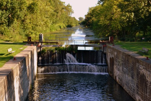 A serene waterway with a small waterfall, surrounded by lush greenery and trees under a clear blue sky.