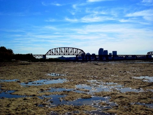 Silhouette of a bridge and city skyline against a blue sky, with a sandy riverbank and reflective water pools in the foreground.