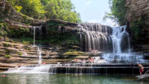 A scenic waterfall cascades over rocky cliffs, with people swimming and relaxing in the clear water below.