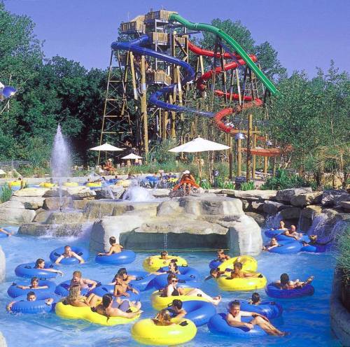 A vibrant water park scene with people floating on inner tubes in a pool, surrounded by slides and greenery.