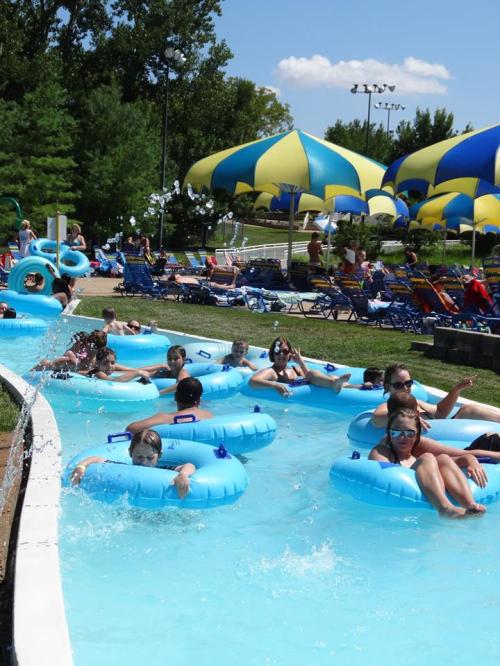 A busy lazy river at a water park with people floating on blue tubes under colorful umbrellas.