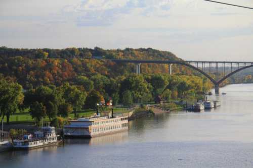 A scenic river view with boats docked, surrounded by trees in autumn colors and a bridge in the background.