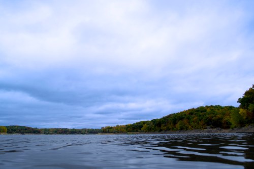 A calm river scene with cloudy skies and trees displaying autumn colors along the shoreline.