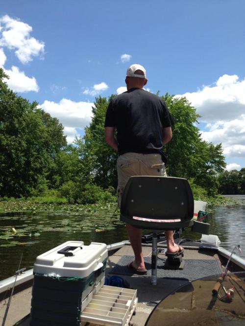 A person fishing from a boat on a calm lake surrounded by lush greenery and blue skies.