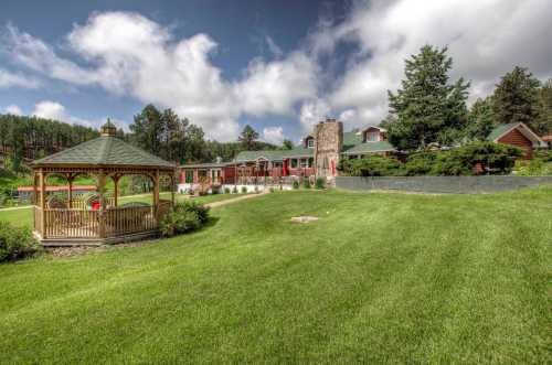 A scenic view of a green lawn with a gazebo, surrounded by trees and a charming building in the background.