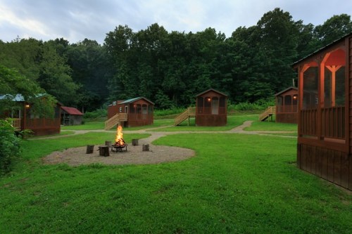 A serene campsite with wooden cabins surrounding a fire pit, set in a lush green area with trees in the background.