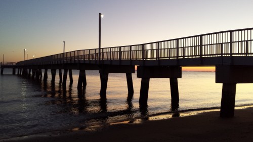 A silhouette of a pier at sunset, with lights illuminating the walkway over calm water.