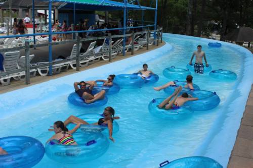 A lazy river with people floating on blue inner tubes, surrounded by a pool area and trees in the background.