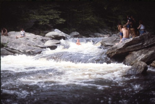 A river with rapids flows between large rocks, while people sit on the banks enjoying the scenery.
