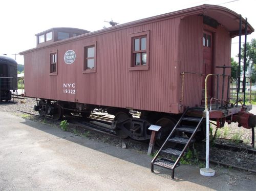 A vintage red caboose with windows and steps, parked on a railway track in a scenic outdoor setting.