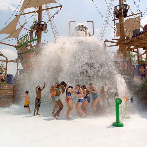 Children and adults enjoy a water splash from a pirate ship at a water park on a sunny day.