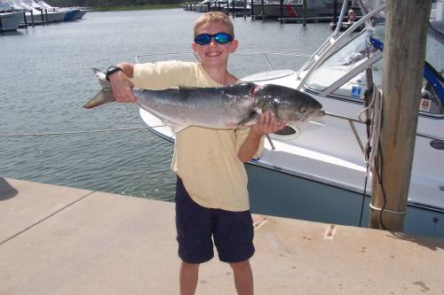 A smiling boy in sunglasses holds a large fish by a marina, with boats and water in the background.
