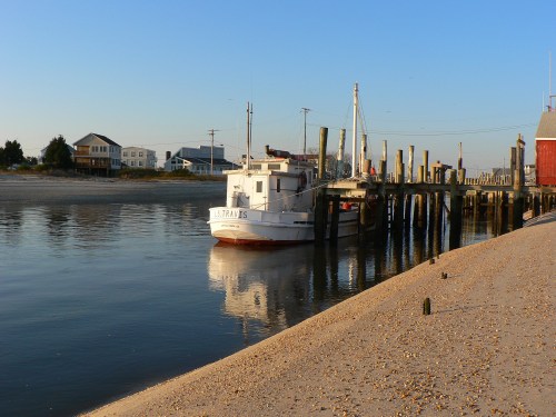 A small boat docked at a pier, reflecting in calm water, with sandy shore and houses in the background under a clear sky.
