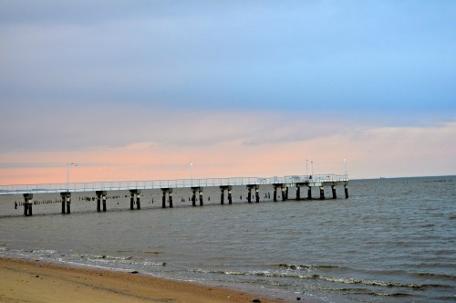 A wooden pier extends into calm waters under a pastel sky, with gentle waves lapping at the sandy shore.