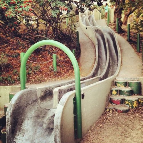 A winding concrete slide in a park, surrounded by greenery and colorful steps leading up to it.