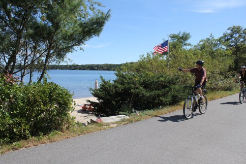 Two cyclists ride along a path by a beach, with trees and an American flag in the background.
