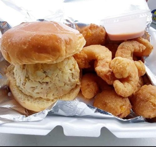 A sandwich with a fluffy bun and crab cake, accompanied by crispy fried shrimp and a small container of sauce.