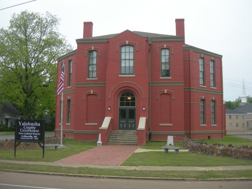 Red brick courthouse with a grand entrance, flanked by trees and a sign for Yalobusha County, Mississippi.