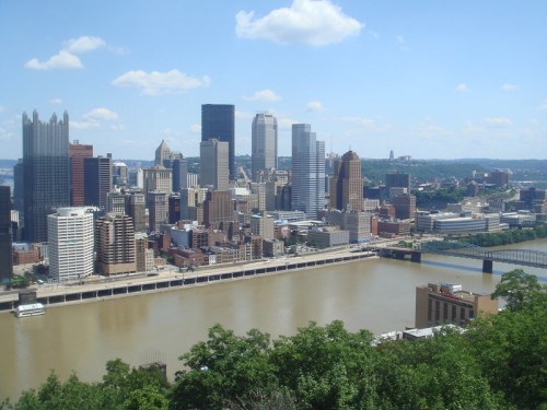 A panoramic view of Pittsburgh's skyline with tall buildings and a river under a clear blue sky.