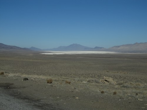 A vast, arid landscape with a salt flat in the distance, surrounded by mountains under a clear blue sky.