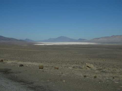 A vast, arid landscape with a salt flat in the distance, surrounded by mountains under a clear blue sky.