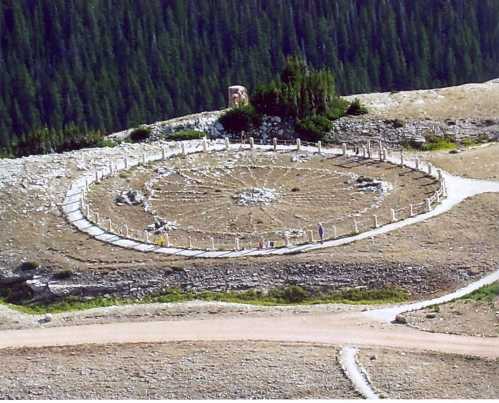 A circular stone structure on a hillside, surrounded by trees, with a path leading to it.