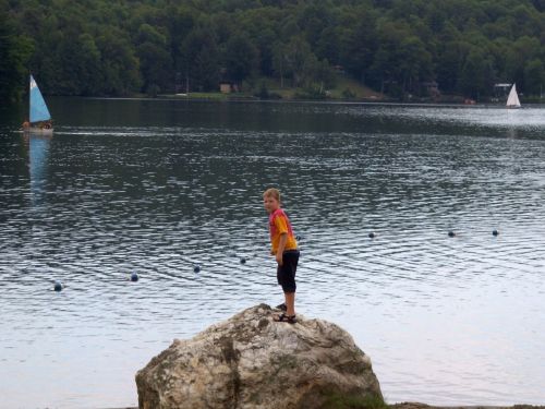 A child stands on a rock by a lake, with sailboats in the background and trees lining the shore.