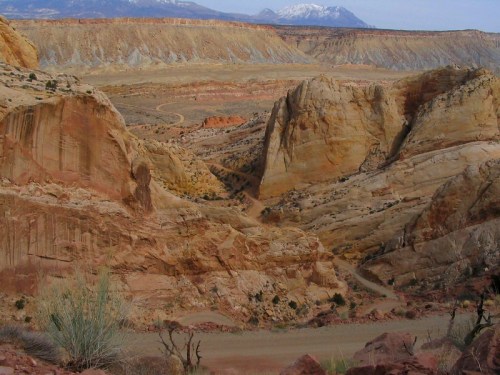 A scenic view of colorful rock formations and a winding path in a desert landscape, with snow-capped mountains in the background.