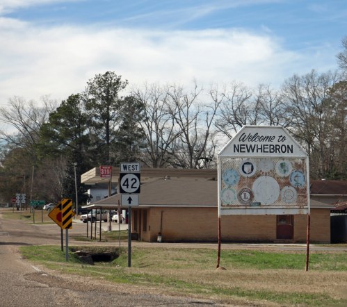 Sign welcoming visitors to New Hebron, with a road sign for Highway 42 and a rural landscape in the background.