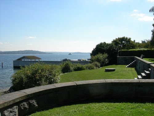 A serene waterfront view with a grassy area, stone wall, and a small boathouse in the distance under a clear blue sky.