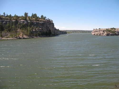 A winding river flows between rocky cliffs under a clear blue sky, surrounded by trees and gentle waves.