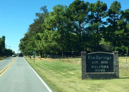 A welcome sign for Elm Springs, established in 1832, alongside a tree-lined road on a sunny day.