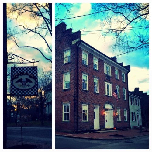 Historic brick building with an American flag, surrounded by trees and a cloudy sky, featuring a sign on the left.