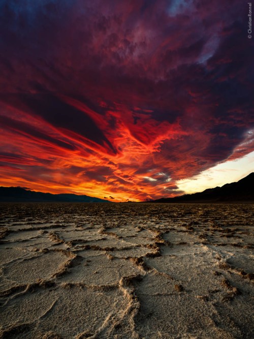 A dramatic sunset over a cracked desert landscape, with vibrant red and orange clouds illuminating the sky.