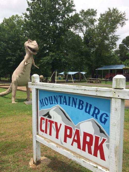 Sign for Mountainburg City Park with a large dinosaur statue in the background, surrounded by trees and playground equipment.