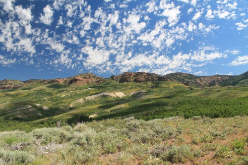 A scenic landscape featuring rolling hills, green vegetation, and a blue sky with fluffy white clouds.