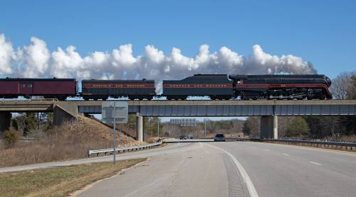 A steam train crosses a bridge over a road, with clouds in the background and vehicles below.