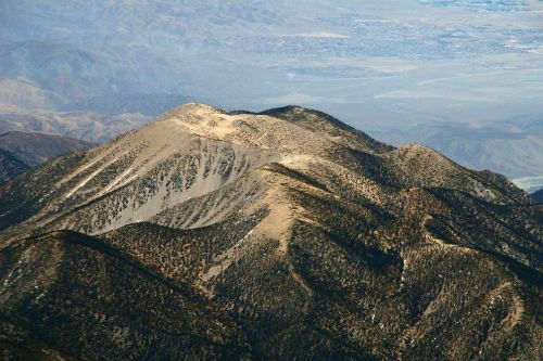Aerial view of a rugged mountain range with rocky peaks and sparse vegetation under a clear sky.