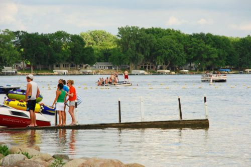 People enjoying a lake day, with boats, a dock, and lush greenery in the background.