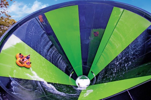 A water slide with a circular, green and black design, featuring riders in a yellow raft descending into the center.