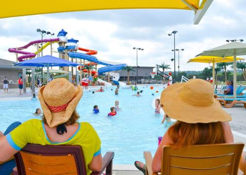 Two women in wide-brimmed hats relax by a pool with colorful water slides, while children play in the water.