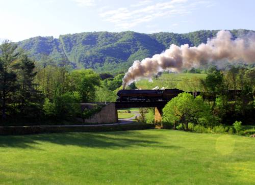 A steam train crosses a bridge, releasing smoke, with lush green hills in the background under a clear blue sky.