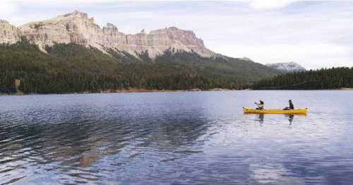 Two people kayak on a calm lake surrounded by mountains and dense forest under a cloudy sky.