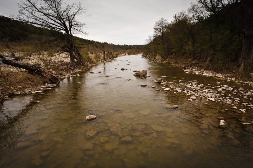 A calm river flows through a rocky landscape, surrounded by trees and a cloudy sky.