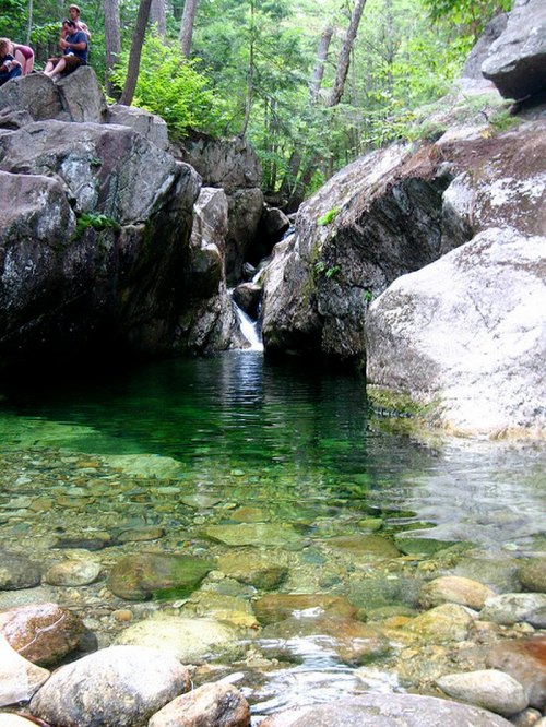 A serene natural pool surrounded by rocks and trees, with a small waterfall in the background and people sitting nearby.