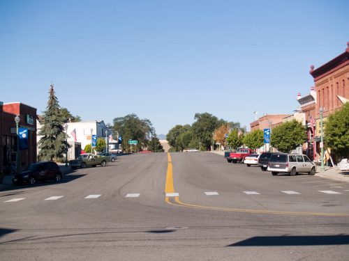 A quiet street in a small town, lined with trees and buildings, under a clear blue sky. Cars parked along the road.