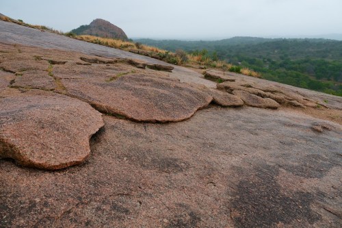 A rocky landscape with smooth, flat granite surfaces and a distant hill under a cloudy sky.