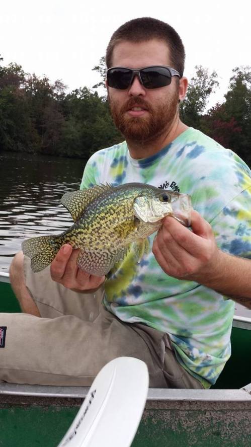 A man in sunglasses holds a large crappie fish while sitting in a boat on a calm lake.