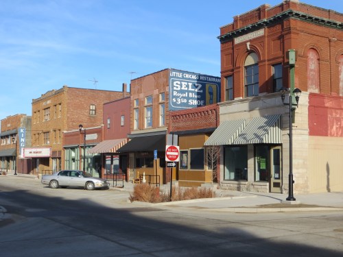 A quiet street scene featuring brick buildings, shops, and a parked car under clear blue skies.